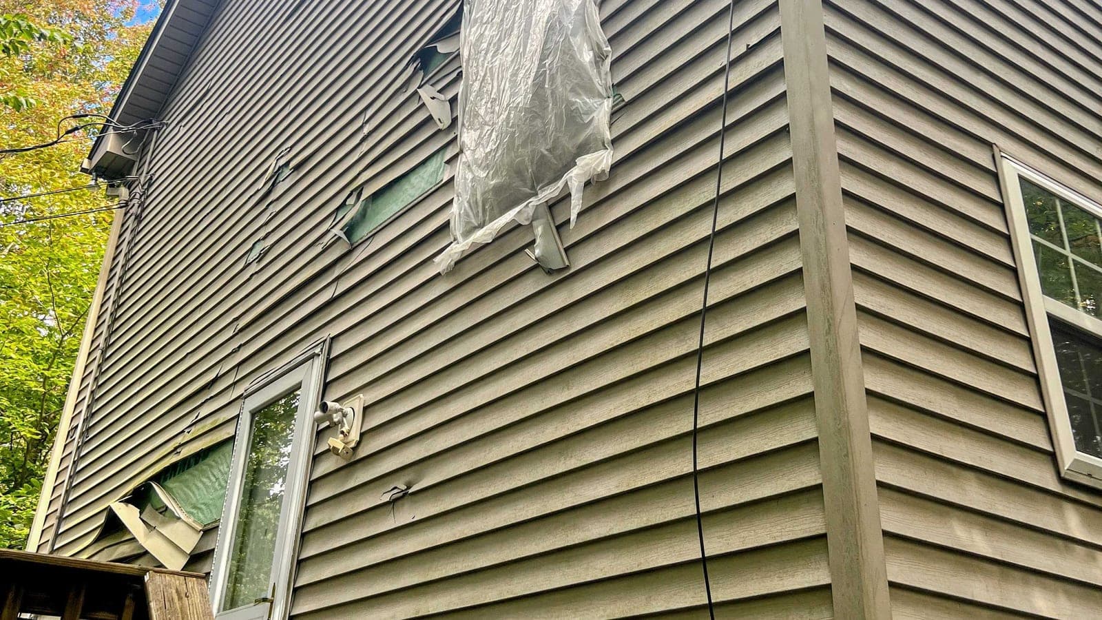 Side of a house with warped, torn vinyl siding and plastic sheeting over a damaged window