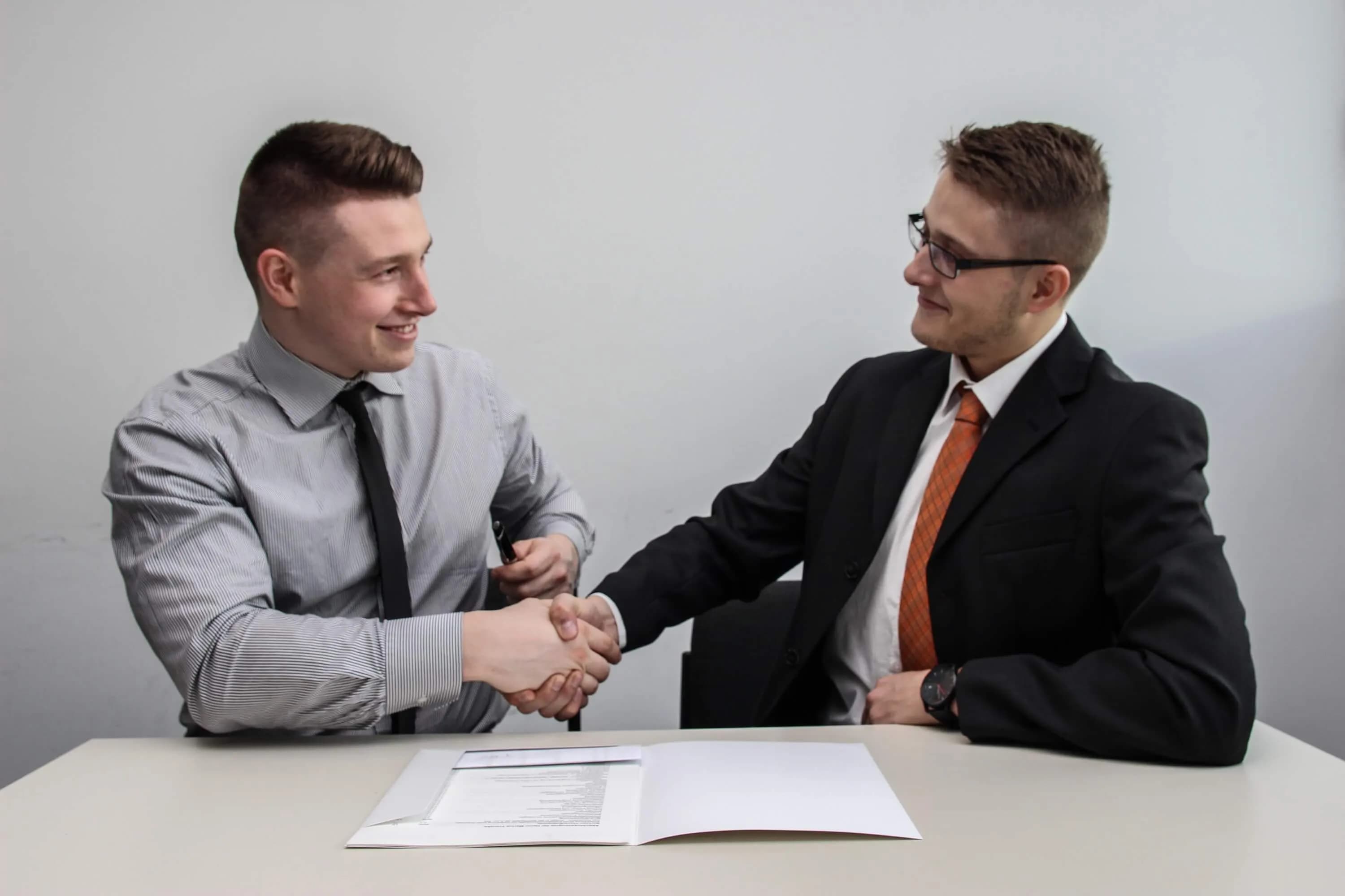 Two men in business clothes shaking hands across a table with an open document folder
