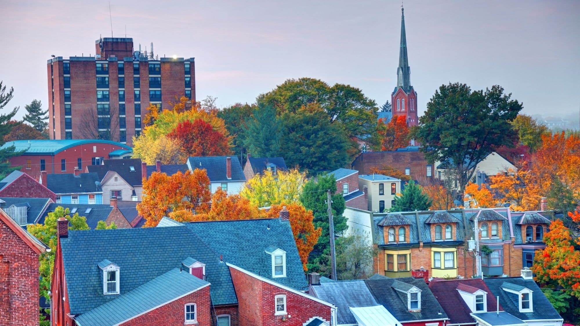Rooftops of brick row houses with dormer windows among orange and yellow autumn trees, with a church spire and a brick high-rise behind